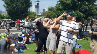 a crowd of people in a city park watch a solar eclipse using safety glasses